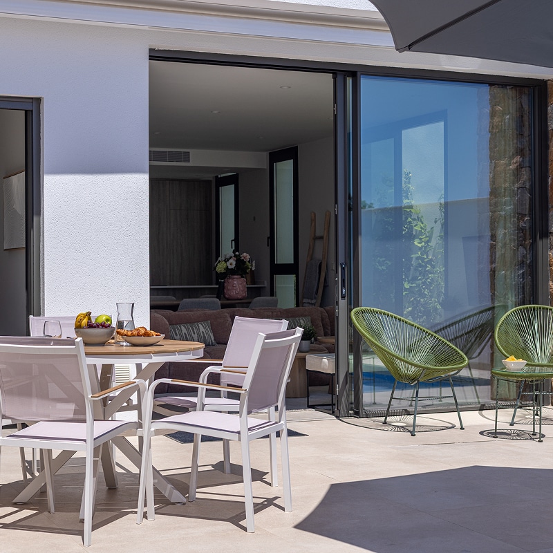 Terrasse avec table à manger, chaises blanches et fauteuils Acapulco verts devant une grande baie vitrée moderne.